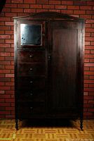 Full view of the tall vintage wooden wardrobe showing the mirror, four drawers, and cabinet door against a brick background.