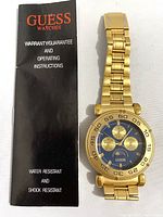 Instruction booklet and Guess watch showing gold-tone metal band, blue dial with subdials and gold markers, watch face and bracelet visible.