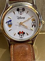 Close-up photo showing the watch face with Mickey, Minnie, Donald Duck, and Goofy characters, gold-tone hands, and hour markers.