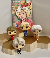 Four Funko Pop Golden Girls dolls displayed on geometric wooden platforms with Betty White Female Force comic book in background.