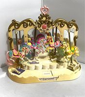 Four pastel-colored carousel horses mounted on brass poles attached to a cream and gold carousel display base with heart top.