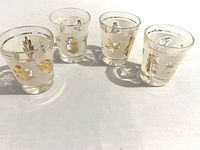 Four vintage clear glass shot glasses with gold-tone foliage designs, shown standing to display motif details and size.