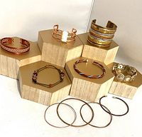 Full set of 10 metal bracelets arranged on hexagonal wooden display mounts, showing copper, brass, and gold finishes, including cuffs, bangles, and a beaded bracelet.