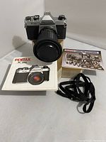 Front view of Pentax K1000 camera with Ozunon 75mm lens mounted, showing camera manual, lenses and accessories booklet, and black strap.