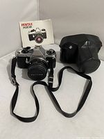Full lot with Pentax ME Super camera, 50mm lens, lens cover, black leather case, neck strap, and instruction manual shown with camera and lens centered and case to the side.