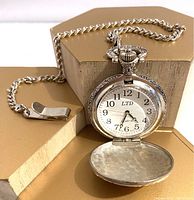 Front view of open pocket watch with white face, black Arabic numerals, and chain laid out to side on tan surface.