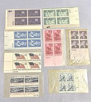 Multiple blocks of four uncanceled US postage stamps in various colors and designs, preserved in wax paper packets and sleeves.