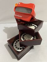 Red View-Master 3D viewer resting on top of the wooden storage box with angled drawers partially open, showing reels inside.