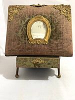 Front view of vintage photo album with pinkish velvet cover, ornate metal corners, oval mirror centerpiece, clasp, and drawer with metal pull on gold-toned legs.