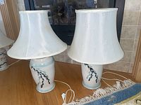 Photo showing pair of Aberle pottery table lamps with mismatched white shades, one bell-shaped and one tapered, placed on wooden surface with fireplace background.