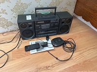 Panasonic boombox with cassette player, radio tuner, two speakers, remote control, power adapter and portable CD player on floor.