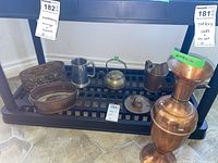 Photo of metal items on black metal shelf including copper watering can, brass kettle, beer mug, plant holders, and copper vase