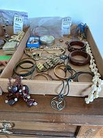 Full lot view showing assorted bracelets, earrings, necklaces, and pendants in a cardboard tray including wood inlaid bangles and a shell necklace.