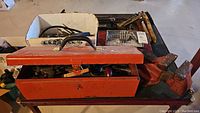 Photo showing orange metal tool box with various hand tools including a bench vice and crow bars on a rolling cart.