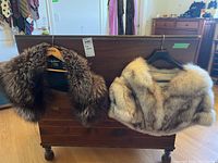 Photo showing two fox fur stoles hanging on wooden furniture, one darker silver fox, one lighter brown fox fur.