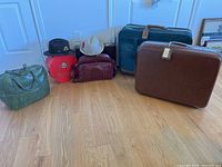 Overview of all items: two hats on top of a red box, green bowling bag, brown briefcase, and two hard-sided suitcases against the wall, showing size and relative scale.