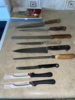 Wide view of all knives and tools in the lot laid on counter: five Henckels knives, two boxed Paderno steak knives, two unboxed Paderno steak knives, sharpening steel and sharpener.
