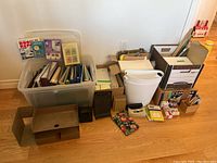 Photo shows a large plastic bin containing assorted binders and folders along with various office supplies including garbage bins, file boxes, rolls of tape, and other smaller items on a hardwood floor against a wall.