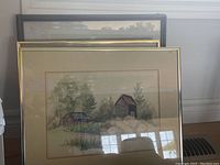 Framed watercolour painting depicting two small barns surrounded by trees. Shows matting and wood frame under glass.