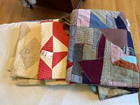 Three folded quilts stacked with the handmade patchwork quilt on top showing varied fabric pieces in different colors like pink, blue, and brown; second quilt in red and white geometric pattern; third quilt in neutral colors