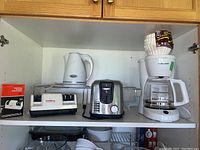 Photo showing Chef's Choice Diamond Hone Sharpener, Black & Decker toaster, Betty Crocker kettle, Black & Decker coffee maker with filters and two metal storage racks inside a cabinet shelf.