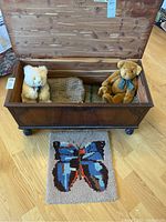 Wooden chest open showing two stuffed teddy bears and two folded mohair blankets inside, with the hand hooked butterfly rug displayed in front on the floor.