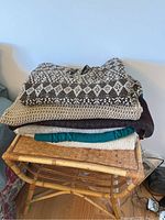 Stack of six folded sweaters in various colors and knit styles: brown and white diamond pattern, beige waffle knit, dark brown, teal green, and off-white, sitting on wicker stool