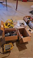 View of whole lot with halogen work light, yellow electrical cable, and multiple boxes containing wiring and electrical parts on a basement floor.