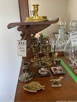 Various brass and silver plate candle holders, brass bells, English Setter figurine, and brass fish-shaped dish arranged on wooden surface with mirror background