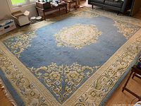 Full view of large rectangular wool area rug with light blue field, central floral medallion, beige border with floral motifs, and fringed edges.