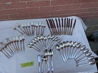 Photo showing the full set of silver plated flatware including forks, knives with dark handles, spoons, teaspoons, and serving utensils displayed on a white surface against a brick wall.