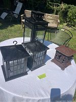 Four hurricane lanterns on an outdoor white table; includes three black metal lanterns and one decorative cast iron lantern with grapevine pattern