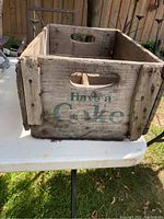 Photo of wooden Coca-Cola crate showing side with text 'Have a Coke' and handle, displaying wear and wood aging.