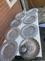 Seven large silver-colored heavy metal plates arranged on a table, displaying raised petal edge designs and textured grid centers, alongside a flower-shaped serving dish with an attached cup.