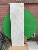 Full front view of rectangular marble slab standing upright against a brick wall and a green circular backdrop, showing overall size and light cream undertones.