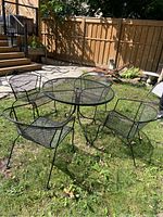 Outdoor photo of black metal patio set with round mesh table and four armchairs on grass near stone patio and wooden fence.