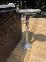 Full side view of the metal pedestal side table on a wooden deck next to outdoor furniture, showing the shape and finish of the table.