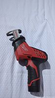 Side view of the Milwaukee M12 HackZall reciprocating saw on a dotted white background, showing red and black design and blade attachment area.