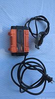 Top view image of the orange and black Motomaster Eliminator battery charger showing the main unit with attached cables and a three-prong power plug.