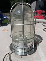 Small marine lighting fixture with protective metal cage and clear glass dome, held in hand to show size and base wiring.
