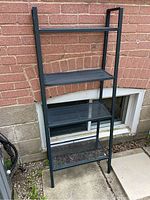 Black metal tiered shelf with four shelves placed outdoors next to a brick wall and basement window. Visible are the different shelf widths and metal frame structure.