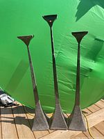 Three metal candle holders of varying heights displayed against a green backdrop on a wooden deck.