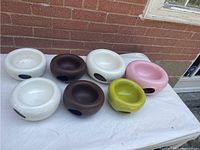 Seven ceramic bowls arranged on a white surface outdoors against a brick wall. Includes two larger white bowls, two brown bowls, one lime green bowl, and one pink bowl.