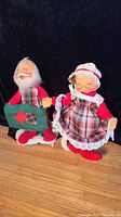 Photo of two vintage Annalee Christmas dolls, Santa and Mrs. Claus, standing on wooden floor with dark backdrop