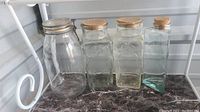 Four apothecary jars on marble surface: 1 round glass jar with a hinged glass lid with metal clasp and rubber washer, 3 rectangular jars with cork stoppers, two have embossed designs with arrows or floral