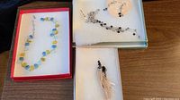 Top view of all four jewelry pieces in presentation boxes: blue and green glass bead necklace with missing clasp in red box, Brighton matching black and silver bead necklace and earrings in green and white boxes, and multi-strand cream bead necklace with black chain.