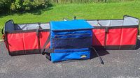 Three car storage items placed on asphalt outdoors: two red foldable compartment bins positioned horizontally and one blue insulated freezer bag in front.