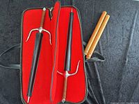 Two practice swords housed in a red-lined carrying case beside a pair of wooden nunchaku on a black surface.