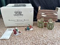 Porcelain Cottage Toy Shop building with two Victorian child figurines in winter attire next to original box showing Heritage Village Collection and Dickens Village Series labeling.