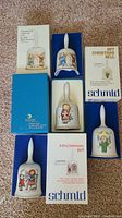 Four vintage ceramic Christmas collector bells each with unique Berta Hummel artwork, displayed with original blue and white Schmid branded boxes labeled with year details.
